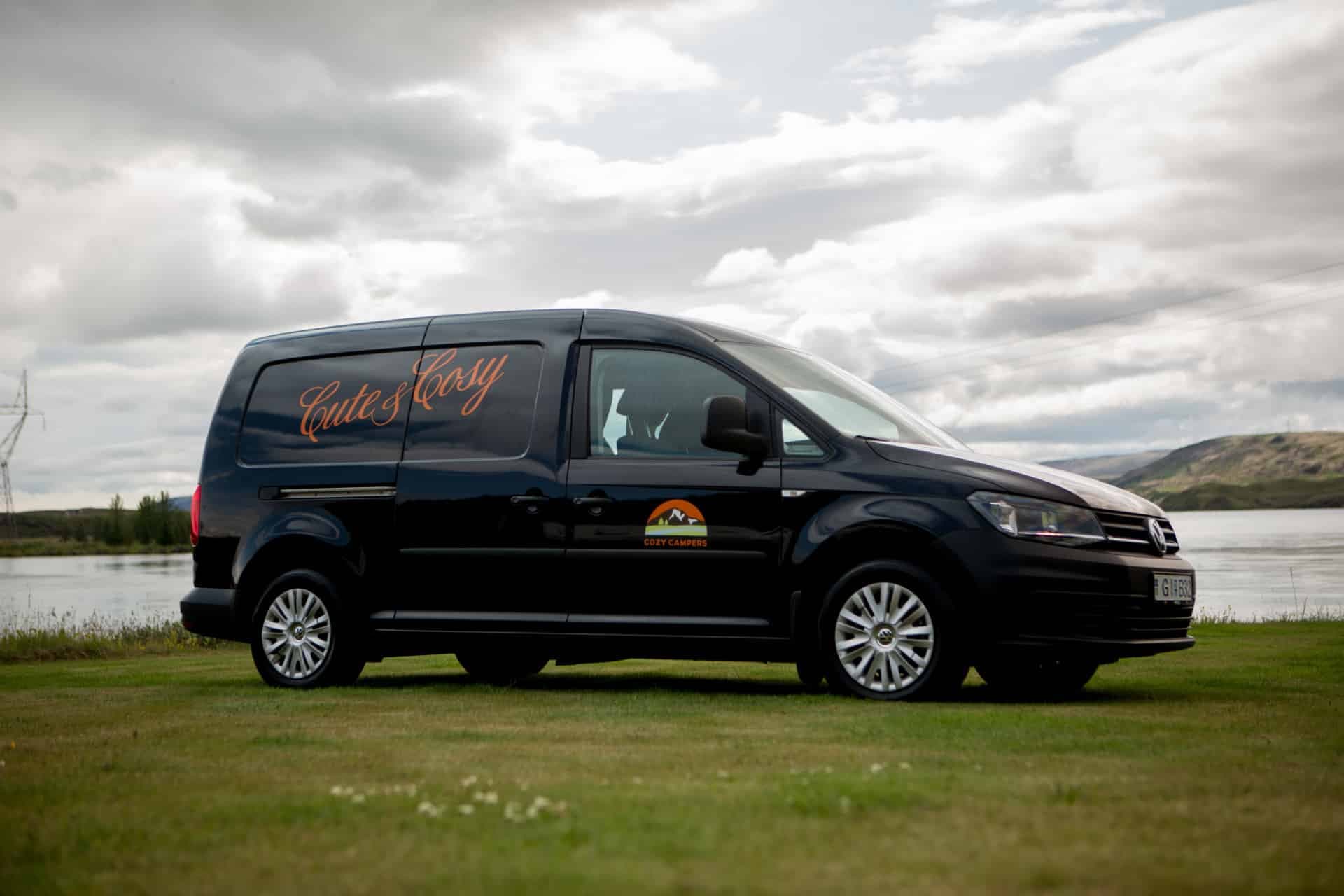 Black VW Caddy camper parked side-on on grass beside a lake, 'Cute & Cosy' script and the Cozy Campers logo on the sliding door