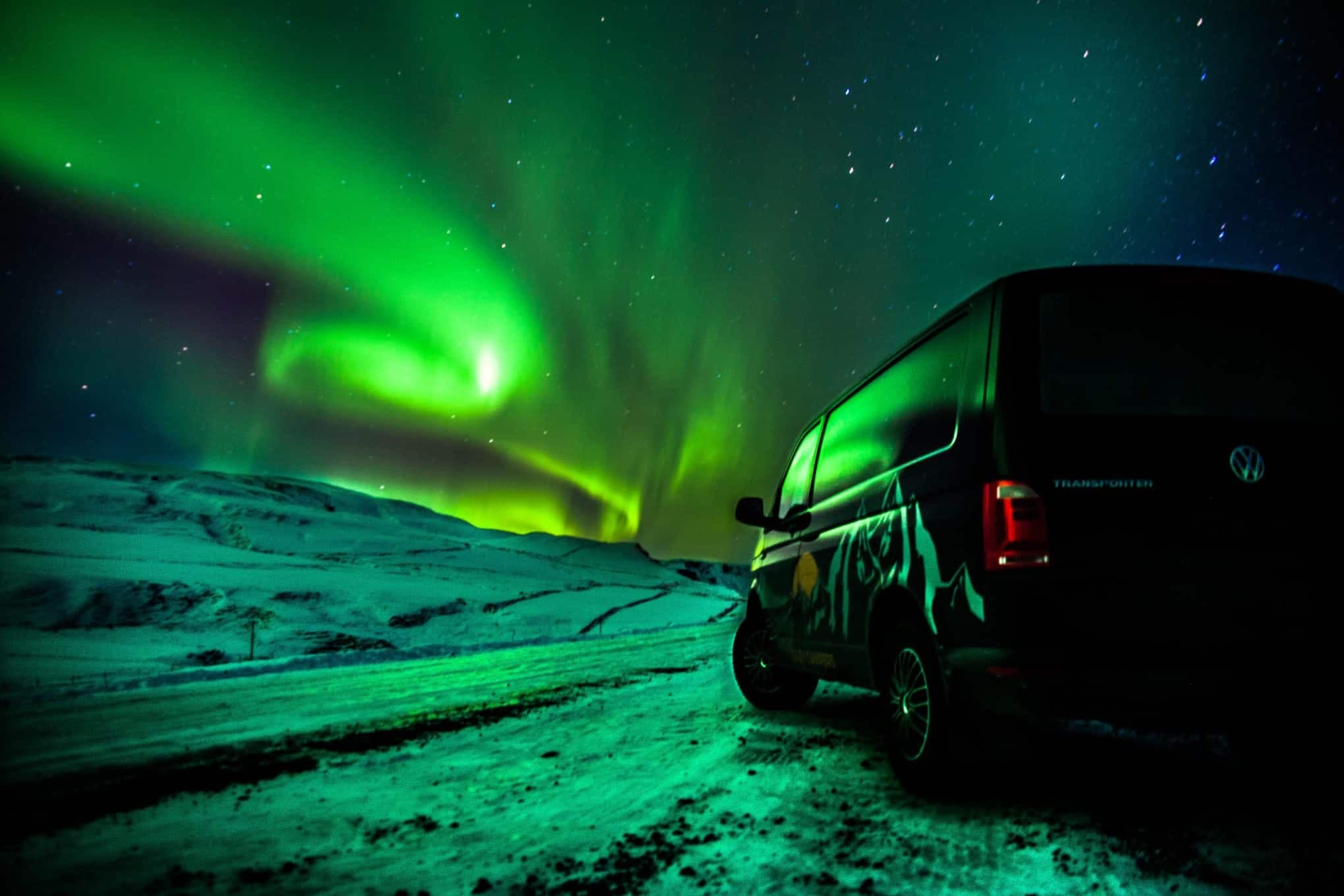Black Volkswagen Transporter camper with Cozy Campers mountain graphic parked on a snow-covered road at night under a green aurora