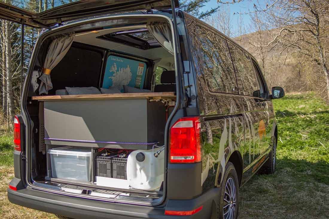 Black VW Transporter camper with rear hatch and sky roof open, bed made up with grey pillows visible inside, parked on grass in front of bare trees and a hillside