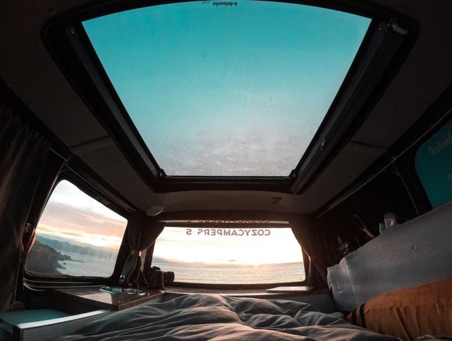 Wide-angle interior shot of the made-up bed with a duvet pulled back, open sky roof above and an ocean sunset visible through the rear window
