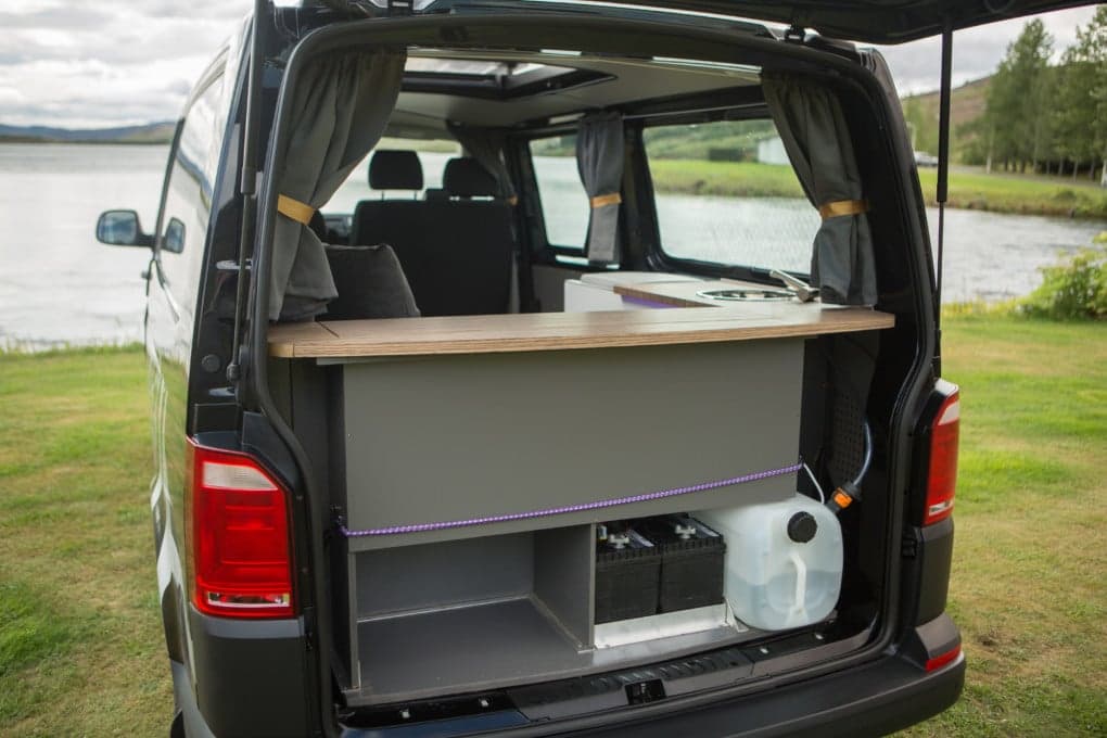 Rear doors open on the Highlander II Budget showing the built-in kitchen counter, water canister and leisure battery, parked beside a lake