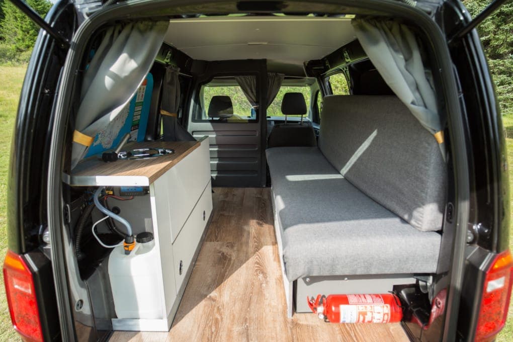 Interior of the VW Caddy looking toward the front seats, with the folded-flat rear bench on the right and a galley counter with sink on the left, no bedding on the bench