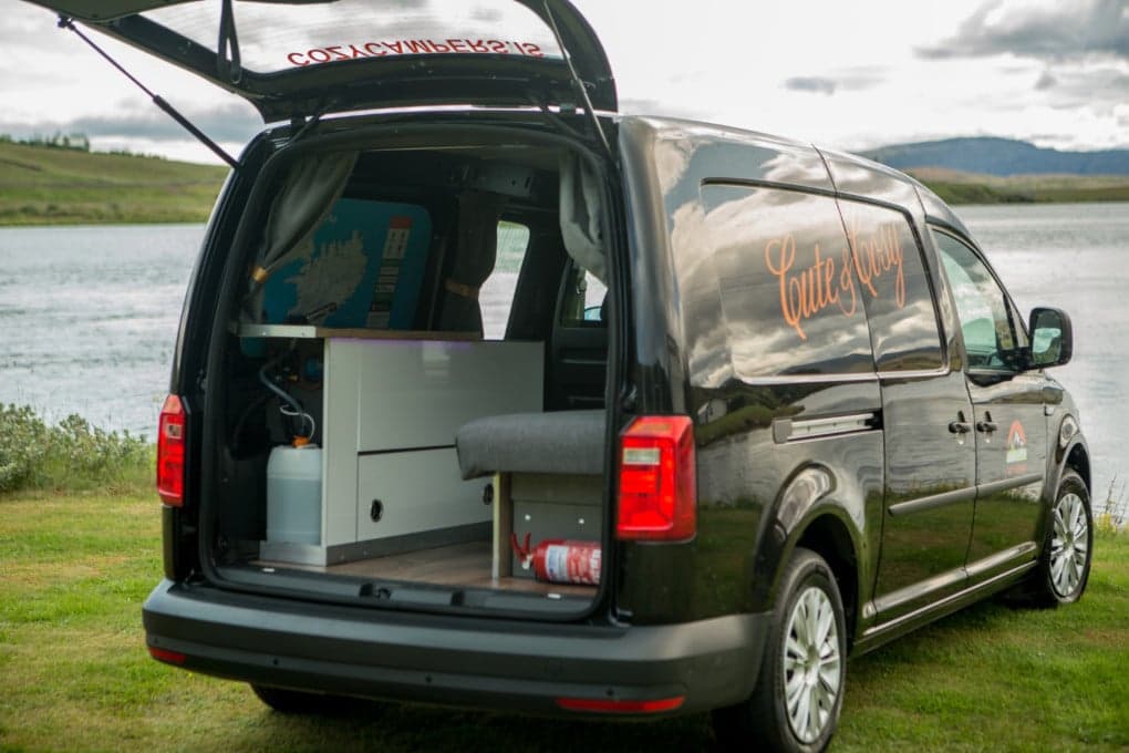 Rear hatch open on the VW Caddy showing the built-in kitchen unit with gas canister, storage drawers and a fire extinguisher strapped beside the folded bench