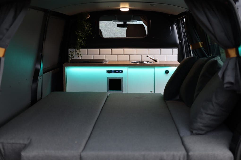 Rear cargo area of the Cozy 3 with the dinette folded flat into a full-width bed, teal LED lighting under the galley units behind