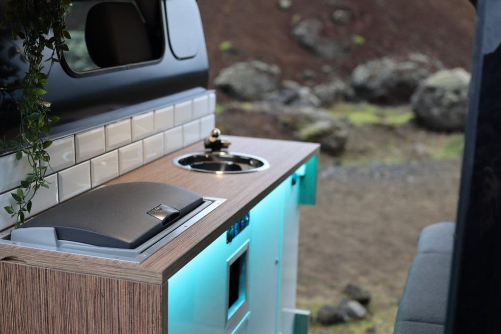 Open side door on the Cozy 3 showing the round sink, cooktop lid and teal-lit cabinet base against a lava-rock backdrop