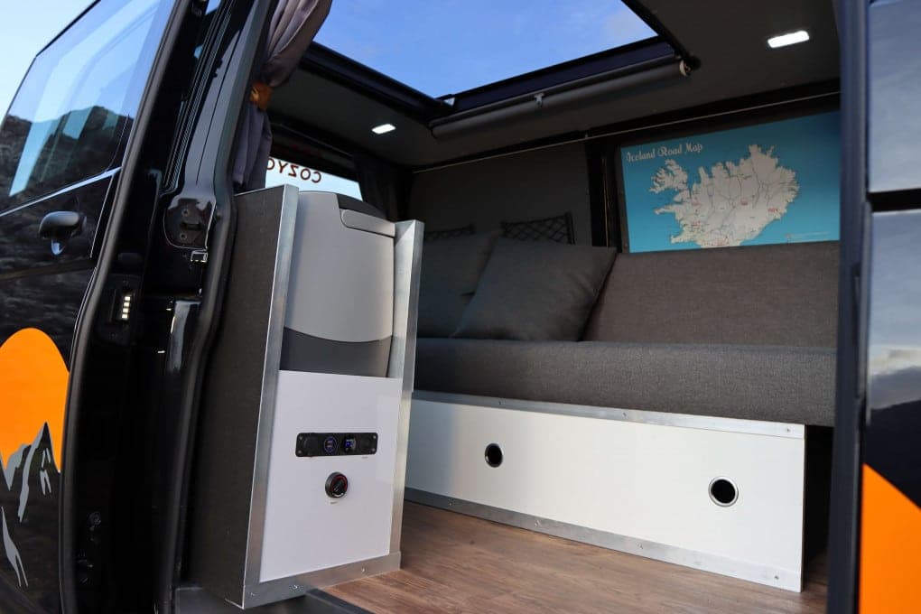 Rear interior of the Highlander I with the side door and sky roof both open, bed made up with grey pillows and an Iceland road map on the wall, heater control panel on the cabinet to the left