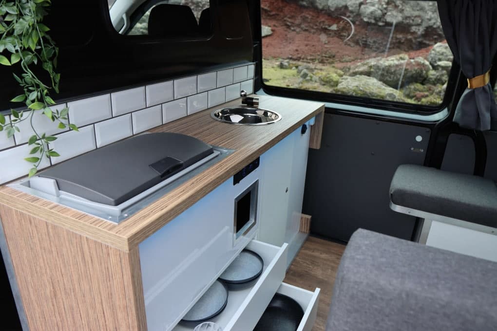 Galley unit with stainless sink, closed stovetop lid, subway-tile backsplash and an open drawer of stacked plates