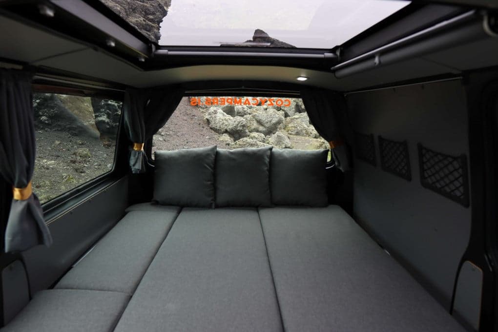 Rear interior of the Highlander III with the full-width bed made up, three pillows against the back window and the sky roof open above