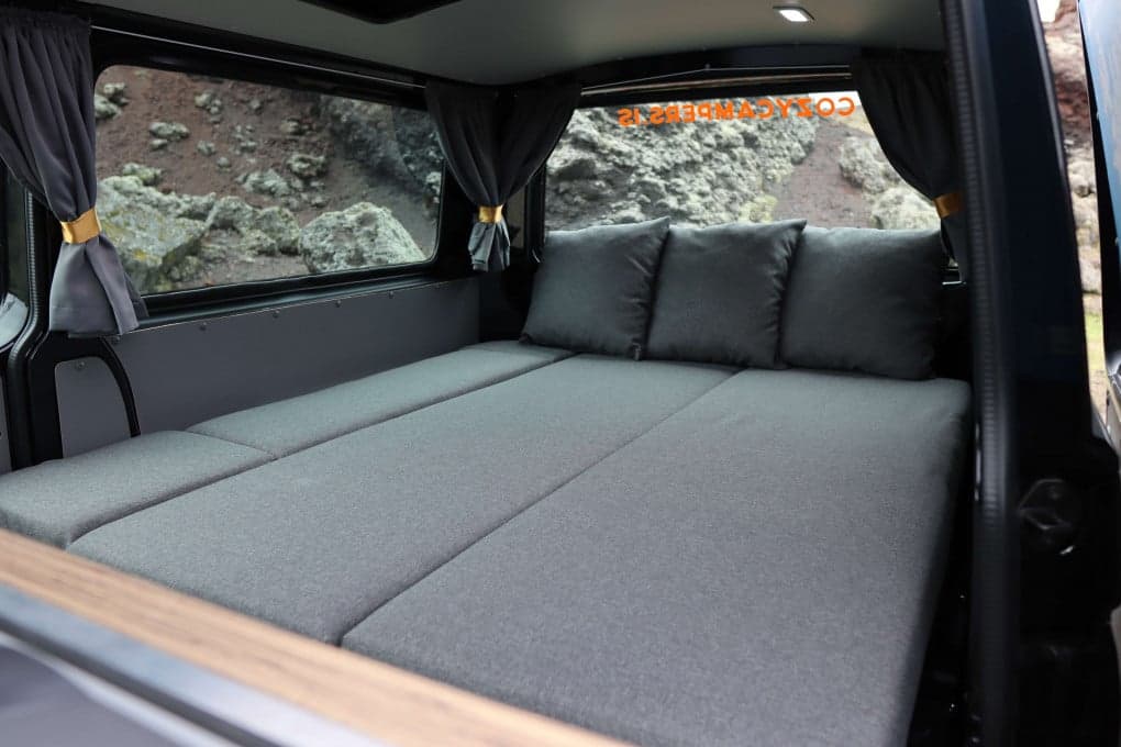 Rear interior with the full-width bed folded flat and made up, three grey pillows against the back window and red volcanic rock visible through the windows
