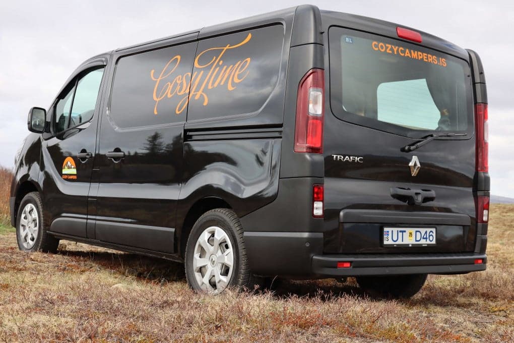 Black Renault Trafic camper viewed from the front three-quarter, Cozy Campers mountain logo on the door, parked on gravel with mountains behind