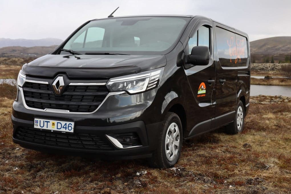 Black Renault Trafic camper parked nose-on on moss-covered lava rock beside a lake, Cozy Campers logo and orange cursive script on the front door