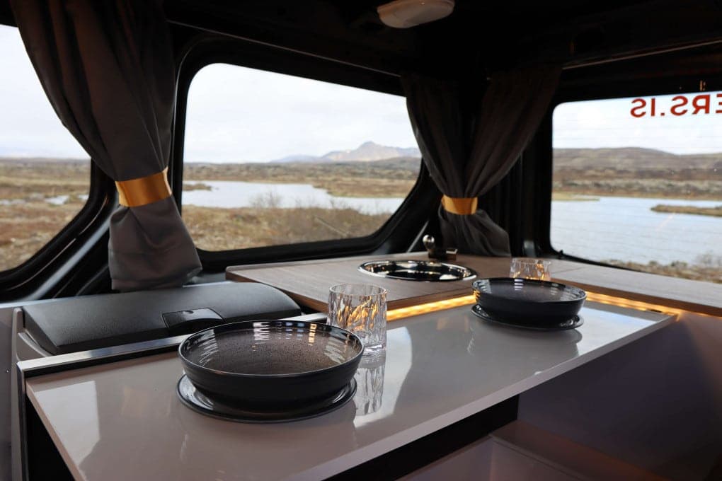 Dinette table set with two plates, a glass and cutlery in the Cozy 2, a lake and mountains visible through the curtained rear windows
