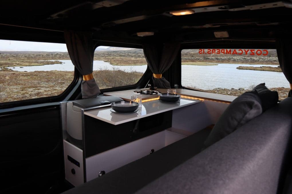 Extendable kitchen table in the Cozy 1 with a built-in two-burner hob, drawers below and two place settings, lake visible through the curtained windows