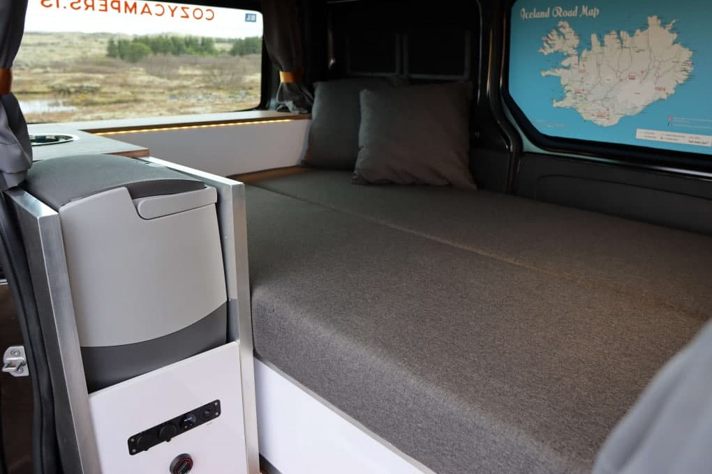 Made-up bed across the rear of the Cozy 1 with two grey pillows, an Iceland road map decal on the back door and a 12V/USB panel on the cabinet