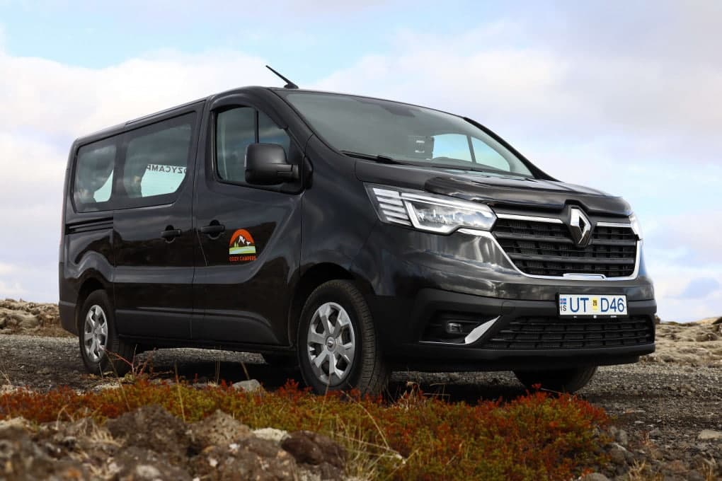 Black Renault Trafic camper viewed from the rear three-quarter, orange 'Cosy Time' script on the sliding door, parked on open grassland