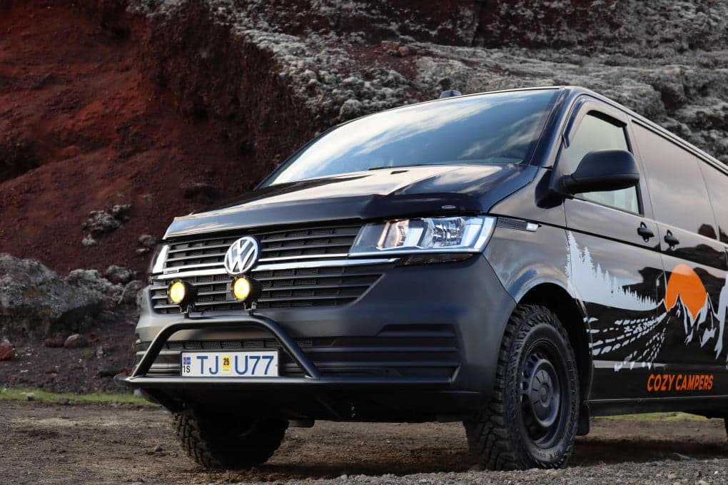 Black VW Transporter camper with off-road tires and a black bull bar with spotlights parked in front of red volcanic rock