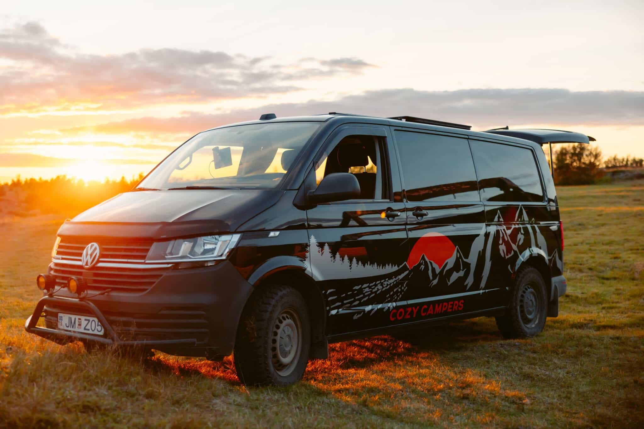 Black Volkswagen Transporter camper with a bull bar, spotlights and a Cozy Campers mountain-and-wolf graphic, parked in a field at sunset