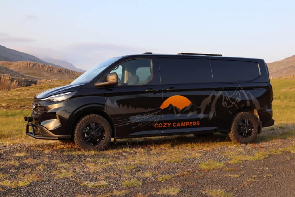 Black Ford Transit Custom camper with Cozy Campers mountain-sunset graphic parked side-on in an Icelandic highland valley, on a lifted 4x4 chassis with off-road tires and a black bull bar