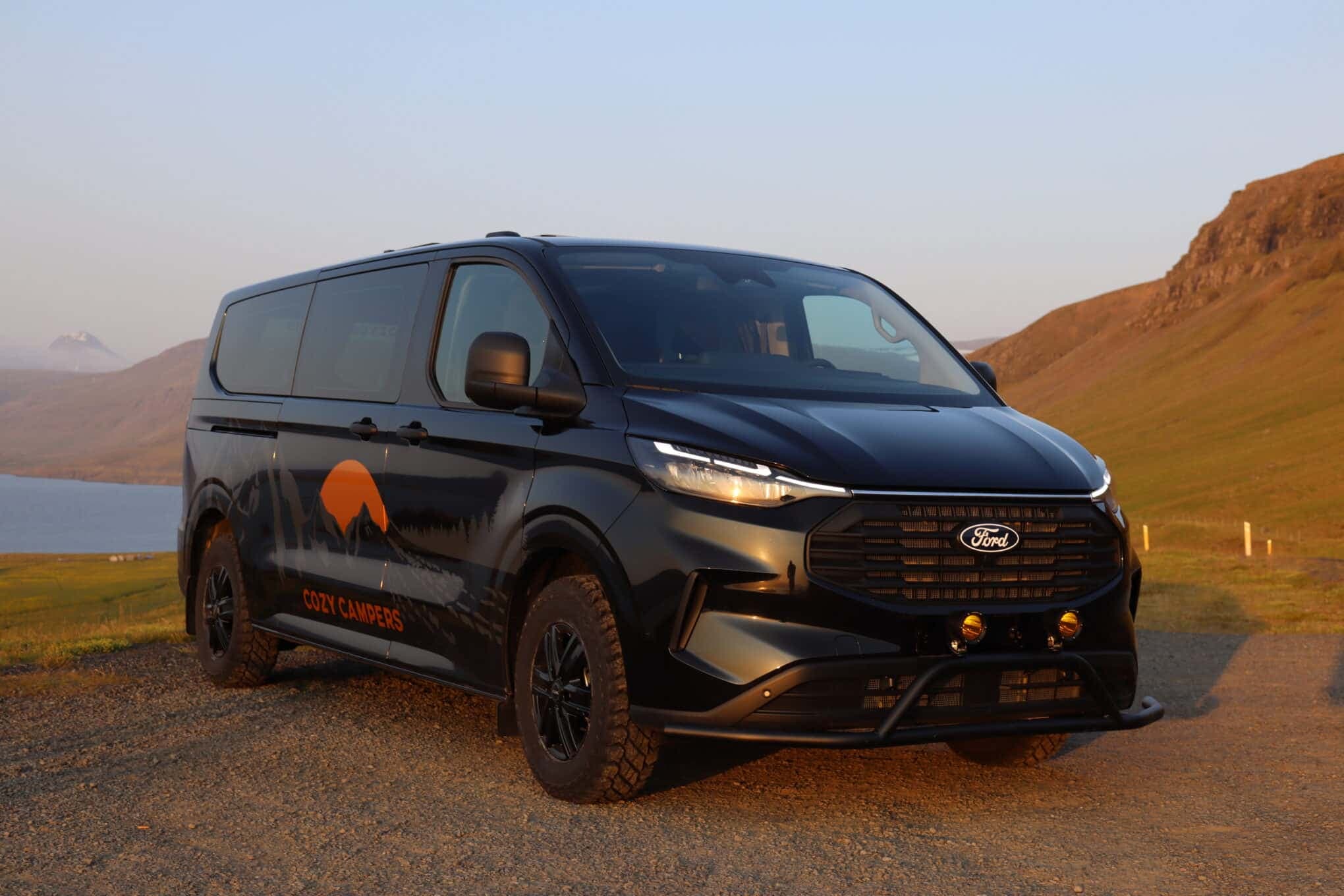 Black Ford Transit Custom camper with lifted suspension and off-road tires parked on a gravel track above a fjord at sunset