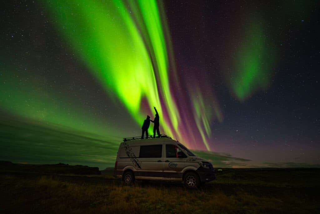 Two people standing on the roof rack of a VW Crafter camper beneath a green aurora