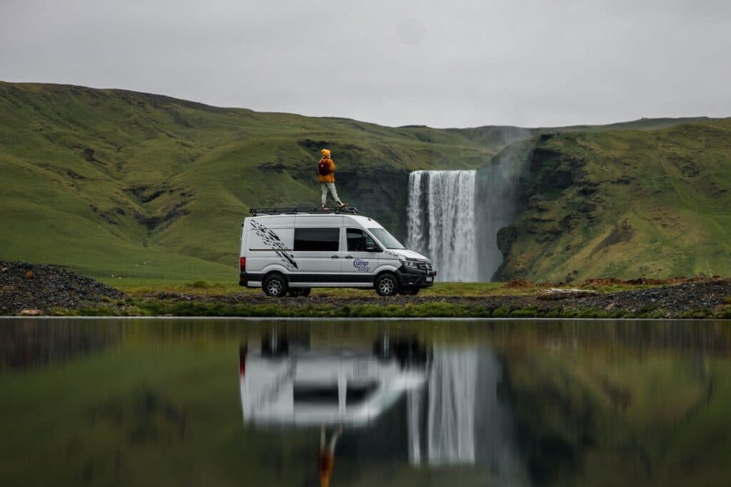 White VW Crafter 4x4 camper with a person standing on its roof rack, mirrored in water in front of Skogafoss