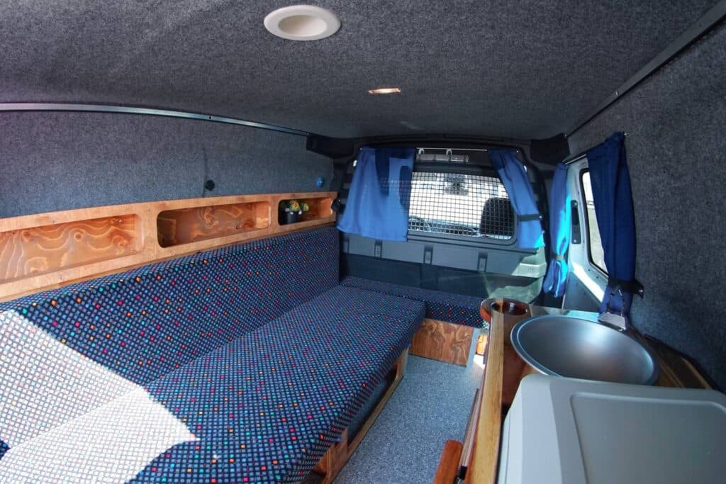 Interior of the Easy Ranger EXP with the bench bed, overhead wooden shelf, sink and blue curtains
