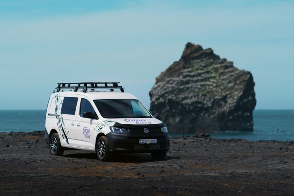 White VW Caddy Maxi 4x4 camper with a roof rack on a black beach in front of a large sea stack