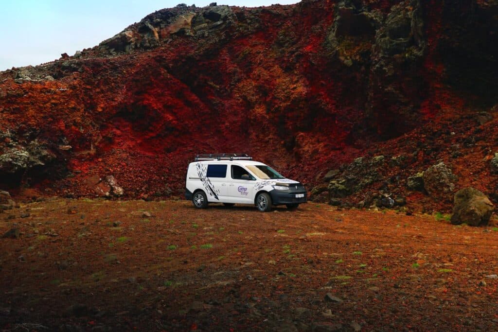 White VW Caddy Maxi 4x4 camper parked below a deep red volcanic crater slope