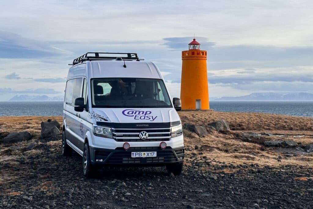 White VW Crafter 4x4 camper with a roof rack parked on lava beside an orange Icelandic lighthouse