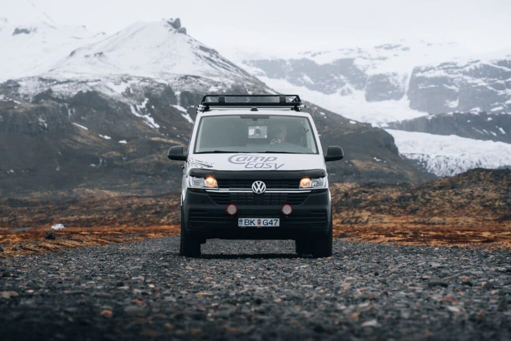 White VW Transporter 4x4 camper with headlights on, parked on black gravel in front of a glacier