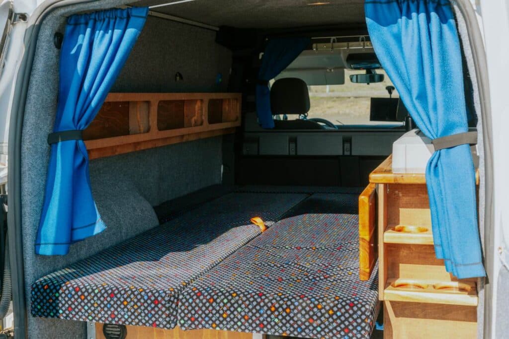 Interior of the Easy Small EXP with the bed base, bright blue curtains and a wooden storage shelf