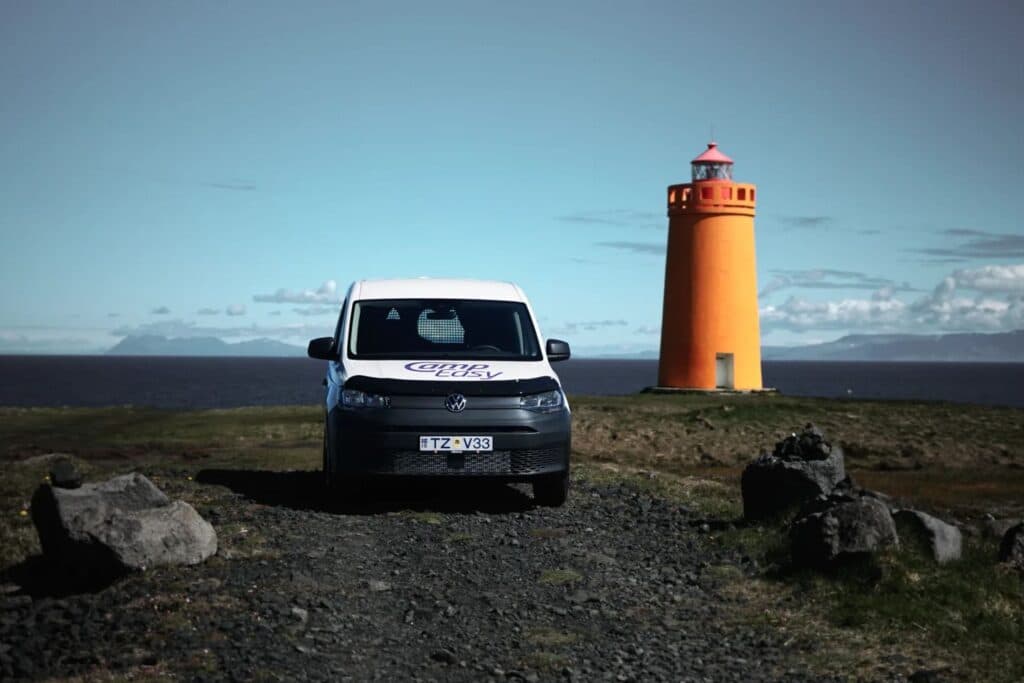 White VW Caddy Maxi camper parked on gravel beside an orange Icelandic lighthouse under a clear sky