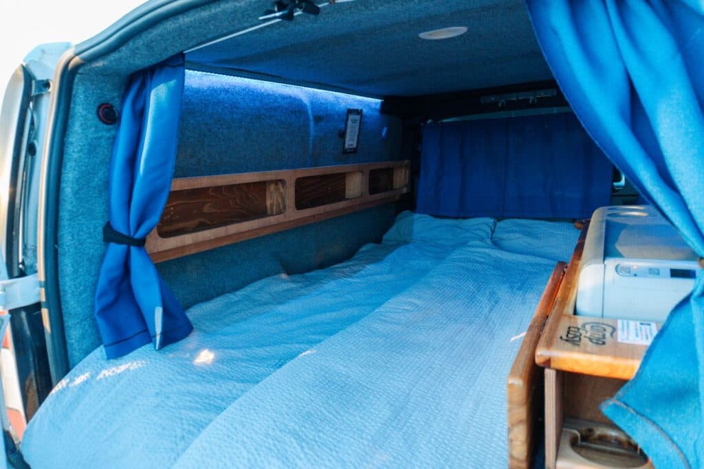 Interior of the Easy Small with the bed made up in blue bedding, blue curtains drawn and a wooden shelf above