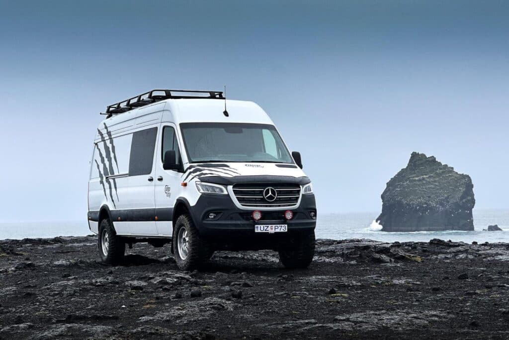 White Mercedes-Benz Sprinter 4x4 camper with a roof rack parked on a black lava beach beside a sea stack