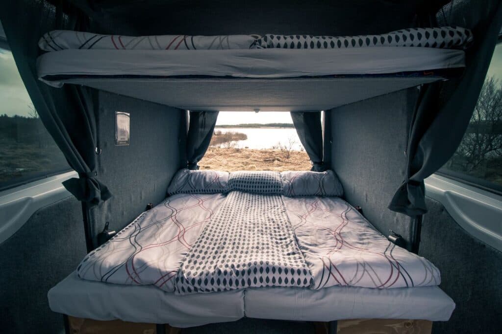 Upper bunk and lower bed made up inside the Easy Big, with the rear windows looking out over a lake