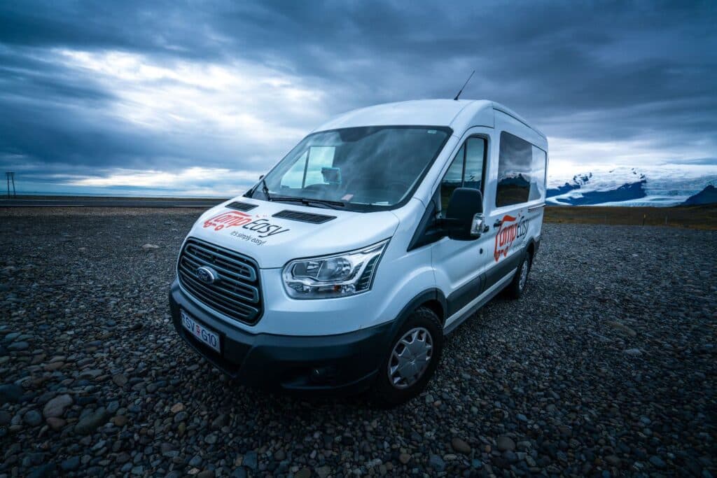 White Ford Transit camper parked on gravel in front of a glacier lagoon under a dark sky