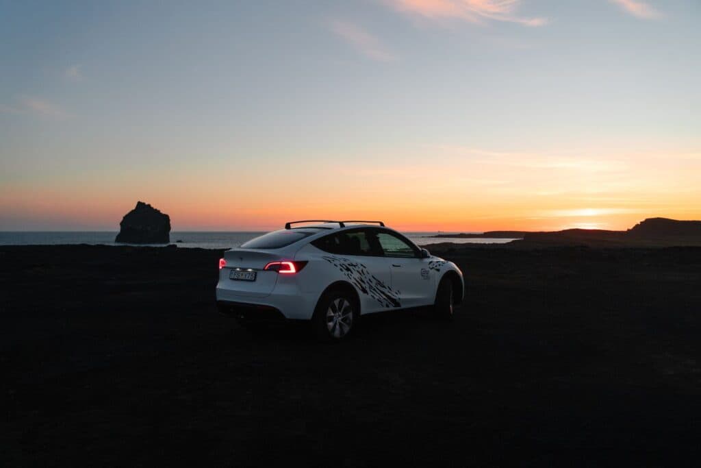White Tesla Model Y parked on black sand at sunset beside a rock formation