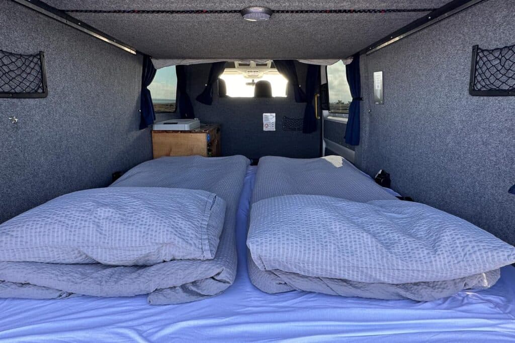 Wide double bed made up with two duvets inside the Easy Viking, blue curtains at the rear windows
