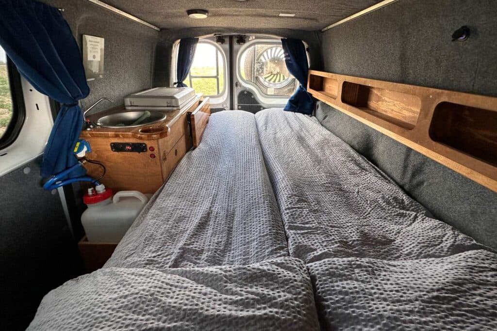 Interior of the Easy Ranger with the bed made up in a grey duvet, a wooden shelf and the sink beyond
