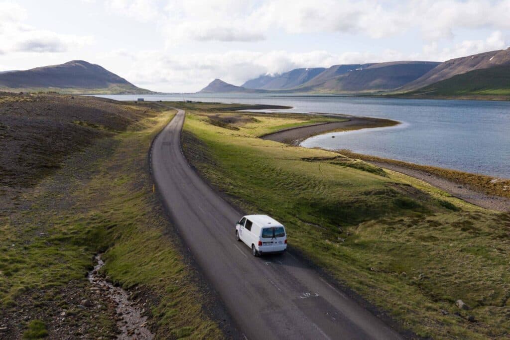 Aerial view of a white VW Transporter camper on a narrow coastal road beside a fjord