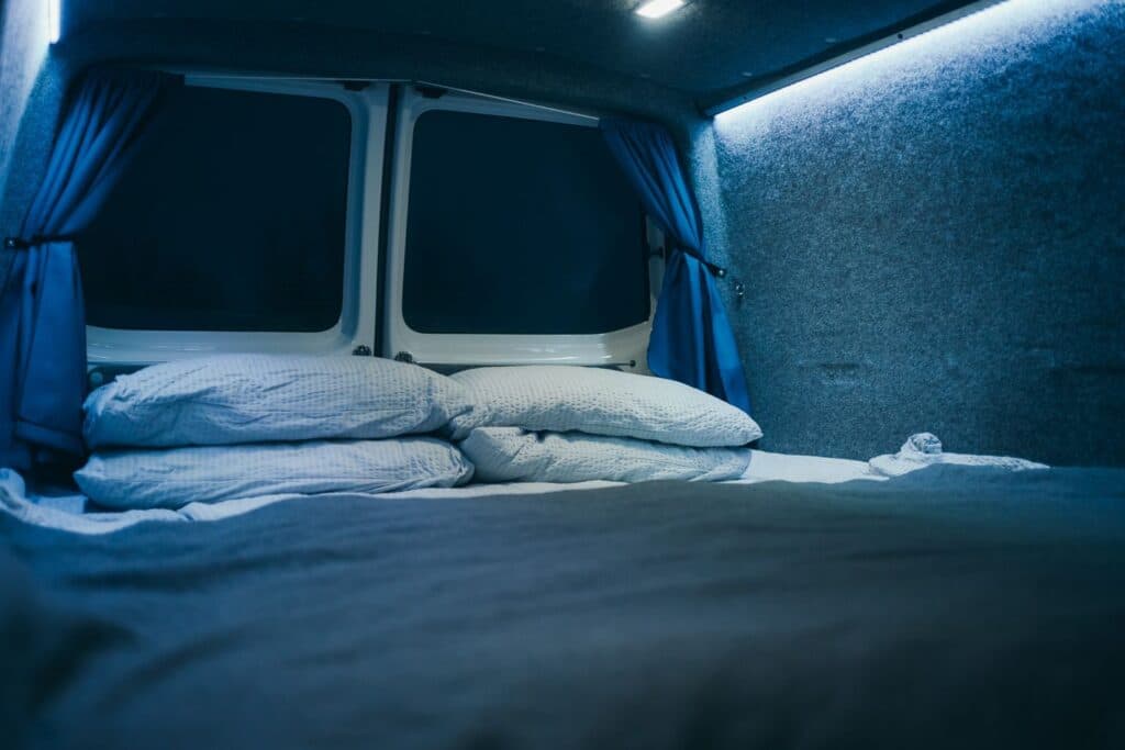 Bed made up with white duvet and pillows inside the Easy Auto EXP, lit by blue interior lighting at night