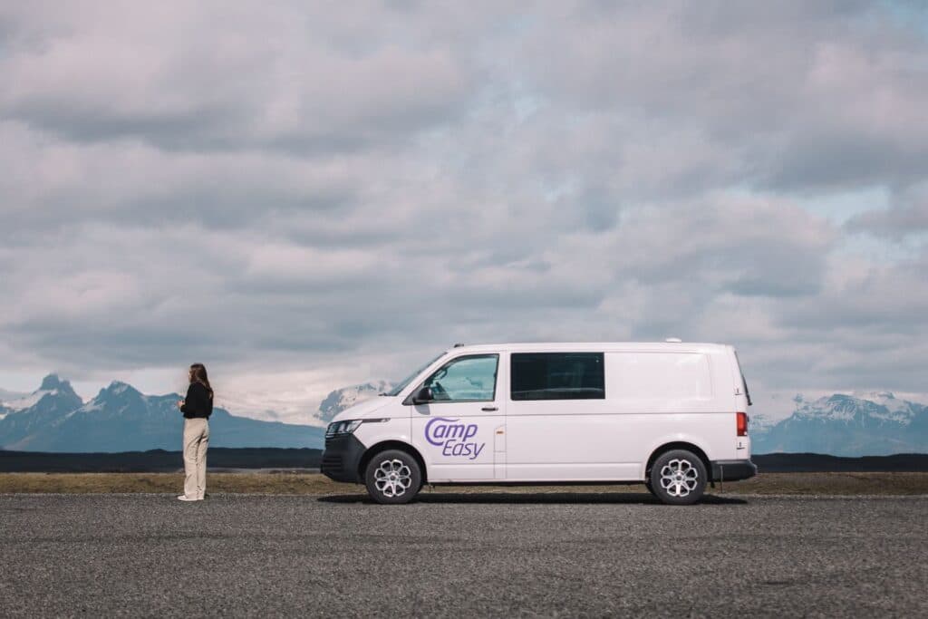 Person standing beside a white VW Transporter camper on a gravel plain with snow-capped mountains behind
