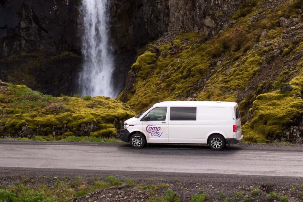 White VW Transporter camper on a road below a mossy cliff with a waterfall behind it