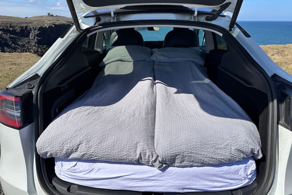 Tailgate of the Tesla camper open with the bed made up in grey bedding, sea and cliffs behind