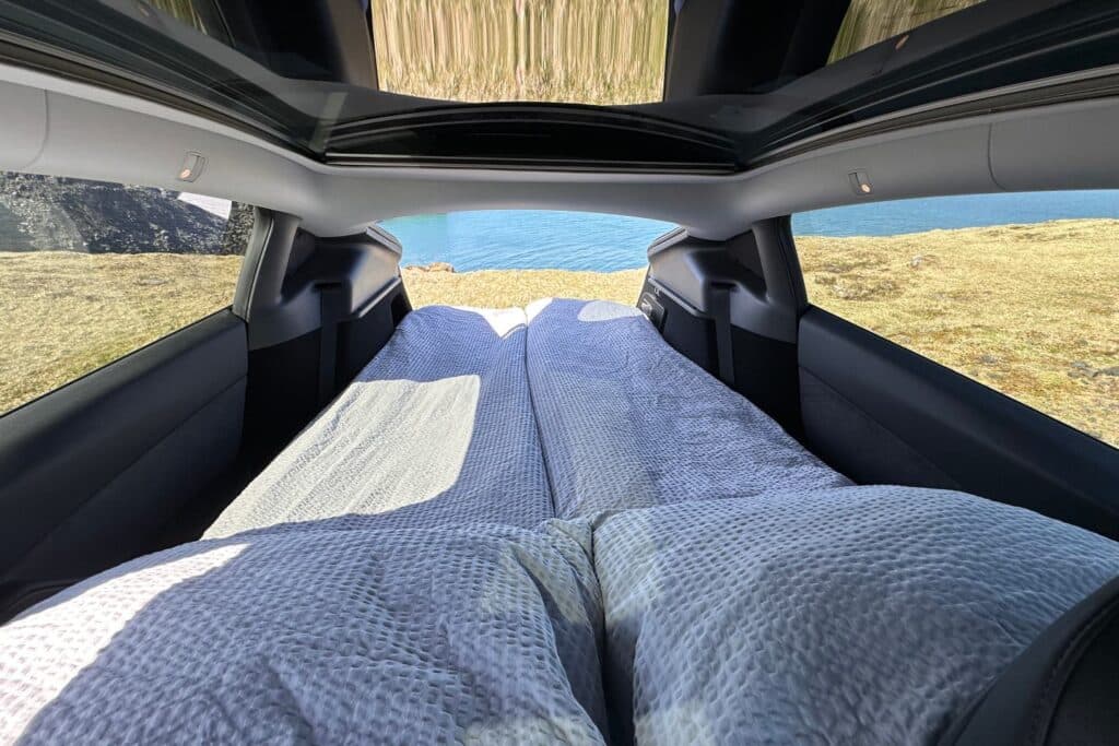 Bed made up inside the Tesla Model Y under the panoramic glass roof, with a coastal view through the tailgate