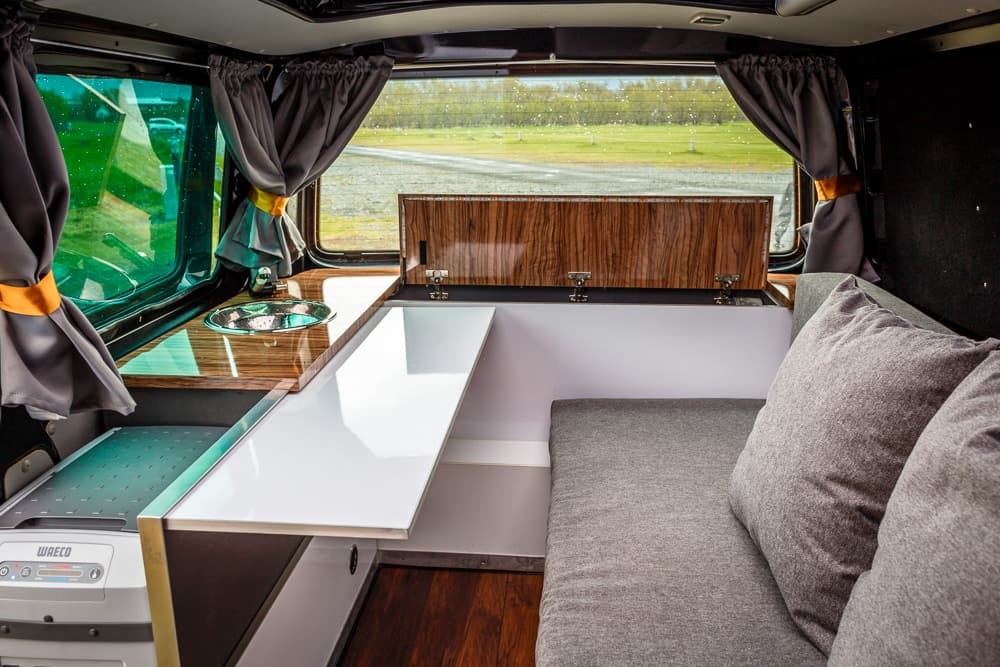 Wood-topped kitchen counter with a round sink and fold-out table in the Highlander I Budget, grey bench cushions with pillows behind and gold-tied curtains at the window